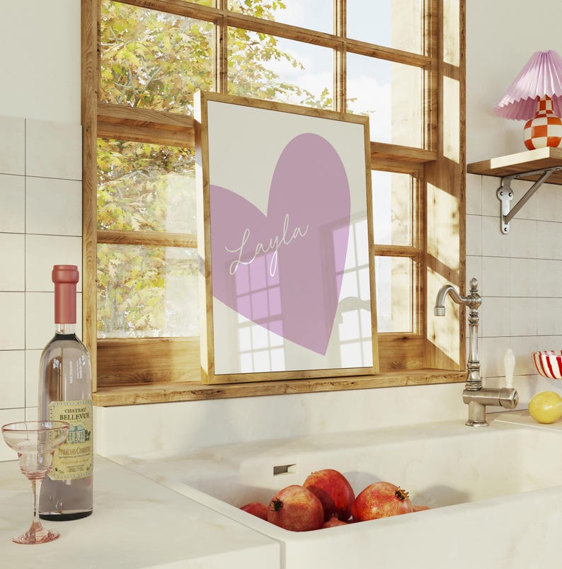 May include: A framed print featuring a large lavender heart with the name "Layla" in cursive script. The artwork is displayed on a windowsill, alongside a bottle of wine, a glass, and pomegranates.