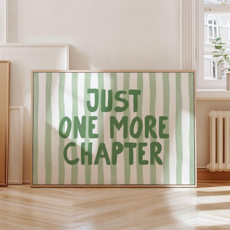 May include: A framed art print with a light wood frame. The artwork features a green and white striped background with the words "JUST ONE MORE CHAPTER" in green, handwritten-style lettering.