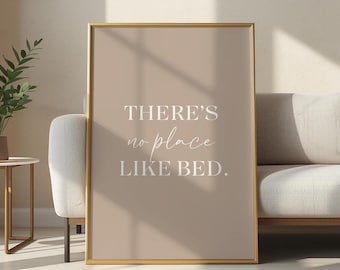 Pode incluir: Uma impressão emoldurada com o texto "THERE'S no place LIKE BED" em branco. A impressão tem um fundo bege e uma moldura dourada. A impressão é exibida em uma sala de estar, ao lado de um sofá e de uma mesa lateral.