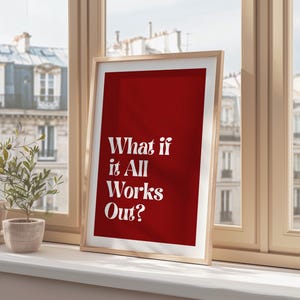 May include: A framed art print with a red background and the white text "What if it All Works Out?". The print is in a light wood frame and is displayed on a windowsill. A small potted plant is visible.
