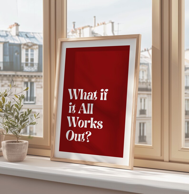 May include: A framed art print with a red background and the white text "What if it All Works Out?". The print is in a light wood frame and is displayed on a windowsill. A small potted plant is visible.