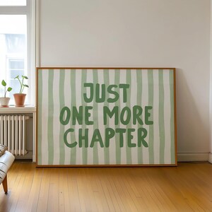 May include: A framed art print with a light brown wooden frame. The artwork features a white background with vertical green stripes and the text "JUST ONE MORE CHAPTER" in green. The print is displayed in a room with a window and hardwood floors.