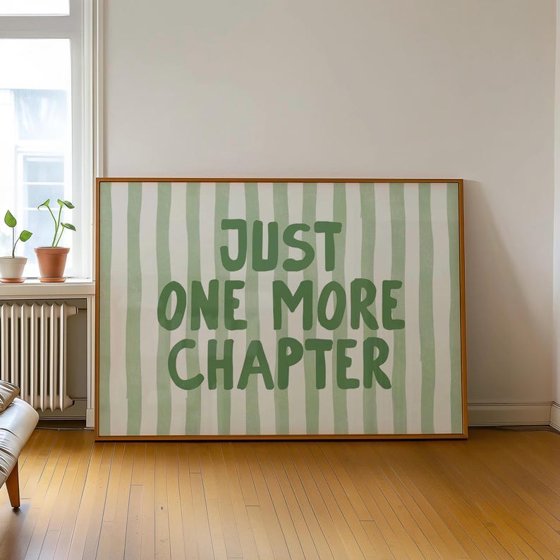 May include: A framed art print with a light brown wooden frame. The artwork features a white background with vertical green stripes and the text "JUST ONE MORE CHAPTER" in green. The print is displayed in a room with a window and hardwood floors.