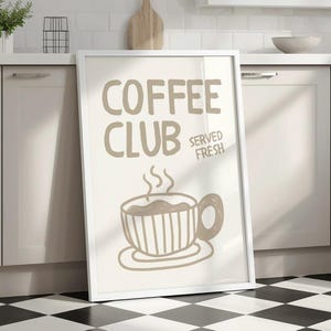May include: A framed print with the words "COFFEE CLUB SERVED FRESH" in a light brown color. The image features a steaming coffee cup illustration. The frame is white, and the background is a neutral color.