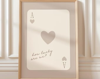 May include: Framed print featuring a playing card, the Ace of Hearts. The card is a soft beige color with a large heart in the center and smaller hearts at the top and bottom. The text "how lucky are we?" is written in a script font.