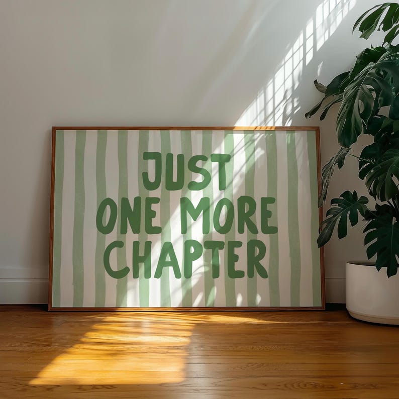 May include: A framed print with the text "JUST ONE MORE CHAPTER" in green letters. The background features vertical green and white stripes. The frame is a warm brown color. A potted plant is visible on the right side of the image.