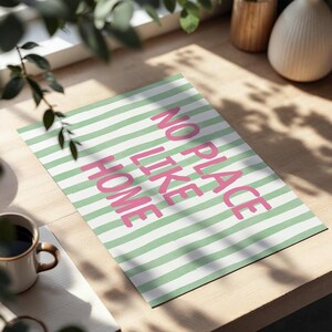 May include: A rectangular art print with a green and white striped background. The words "NO PLACE LIKE HOME" are printed in pink. The print is displayed on a light-coloured wooden surface, with a coffee mug and greenery in the foreground.