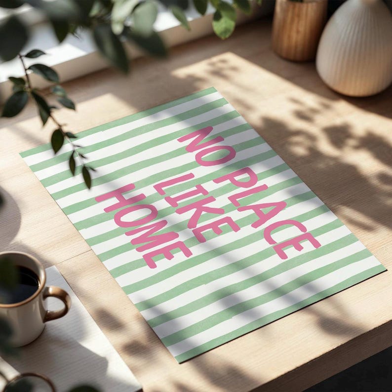 May include: A rectangular art print with a green and white striped background. The words "NO PLACE LIKE HOME" are printed in pink. The print is displayed on a light-coloured wooden surface, with a coffee mug and greenery in the foreground.