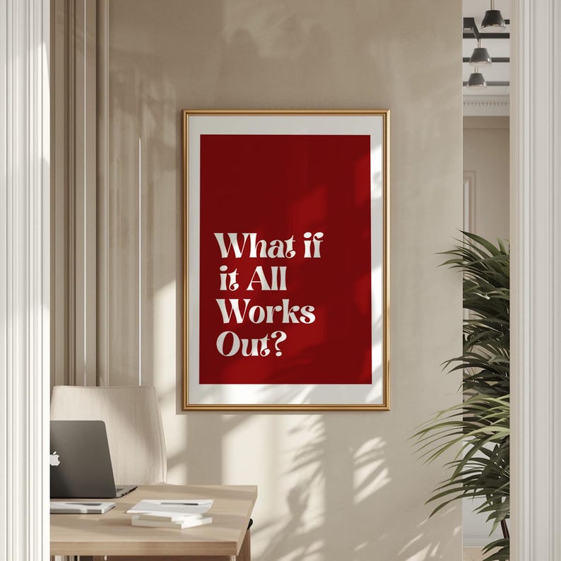 May include: A framed print with a gold border and a deep red background. The white text reads "What if it All Works Out?" The print is hanging on a beige wall, with a desk and laptop visible in the foreground.
