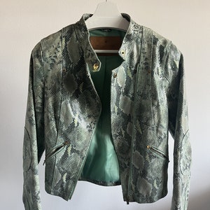 Może przedstawiać: Jasnozielona kurtka ze wzorem skóry węża, stójką i złotymi zamkami. Kurtka ma dopasowany krój i zieloną podszewkę. Nazwa marki "Gocce di Mare" jest widoczna na brązowej skórzanej naszywce.