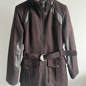 May include: A dark brown coat with black leather accents on the shoulders and sleeves. The coat features a high collar, a full-length zipper, a belt with a silver buckle, and two front pockets. The coat is hanging on a white hanger.
