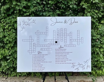 May include: A white sign with a crossword puzzle and the names "Jamie & Dan" in cursive. The crossword has clues related to the couple's life. Decorative floral elements are in the corners. The sign is on a black easel.
