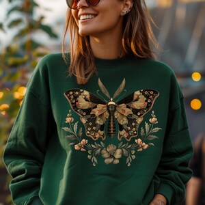 Op de afbeelding: Een donkergroen sweatshirt met een bloemenpatroon met een grote vlinder in het midden. De vlinder is versierd met gouden en zwarte accenten.