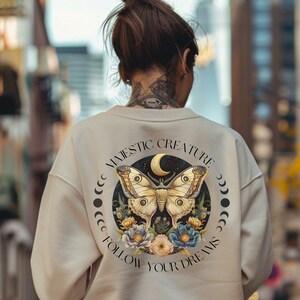 Peut inclure: Sweat-shirt crème avec un motif circulaire au dos. Le motif représente un papillon, une lune, des fleurs et le texte "MAJESTIC CREATURE FOLLOW YOUR DREAMS". Le design est en beige, bleu et rose.