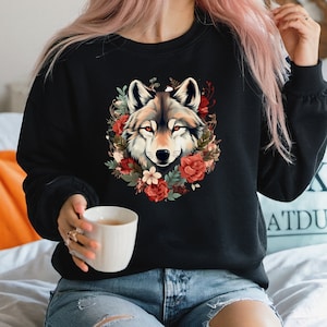 May include: Black sweatshirt featuring a detailed wolf face surrounded by a floral wreath of red roses and greenery. The wolf has gray and brown fur with orange eyes. The sweatshirt is a comfortable, casual style.