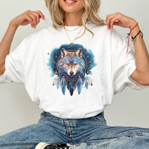 Könnte beinhalten: Weißes T-Shirt mit einem blauen und goldenen Wolf-Traumfänger-Design. Die Augen des Wolfes sind blau und der Traumfänger hat Federn.