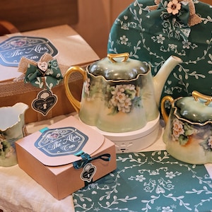 May include: A tea set with a teapot, sugar bowl, creamer, and a tea cozy. The set is painted with white flowers and gold accents. The set is displayed with a gift box and a patterned green fabric.