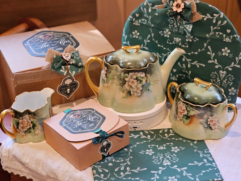 May include: A tea set with a teapot, sugar bowl, creamer, and a tea cozy. The set is painted with white flowers and gold accents. The set is displayed with a gift box and a patterned green fabric.