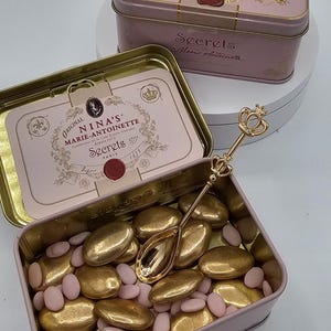 May include: A pink and gold tin box filled with gold and pink candy. The box has a gold crown and the text "ORIGINAL NINA'S MARIE-ANTOINETTE Faveur de la Cour Royale Francaise depuis 1672 Secrets 777 PARIS" on the lid. The box is open and a gold spoon with a crown on the handle is inside.