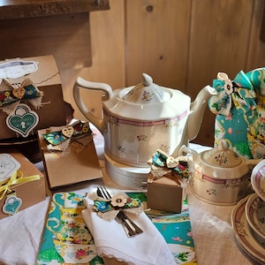 May include: A vintage tea party setting with a white teapot with floral accents, a sugar bowl, a teacup and saucer, and several small brown boxes with burlap bows and floral accents. The boxes are labeled "Tea Party" and "The Alps Tea".