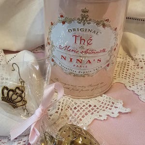 Op de afbeelding: Een roze theedoos met gouden rand en de tekst "ORIGINAL Th&eacute; de Marie Antoinette NINA'S PARIS Versailles depuis 1672". De doos staat op een witte kanttafelkleed. Een gouden hartvormige thee-ei is in een doorzichtige plastic zak met een roze lint.