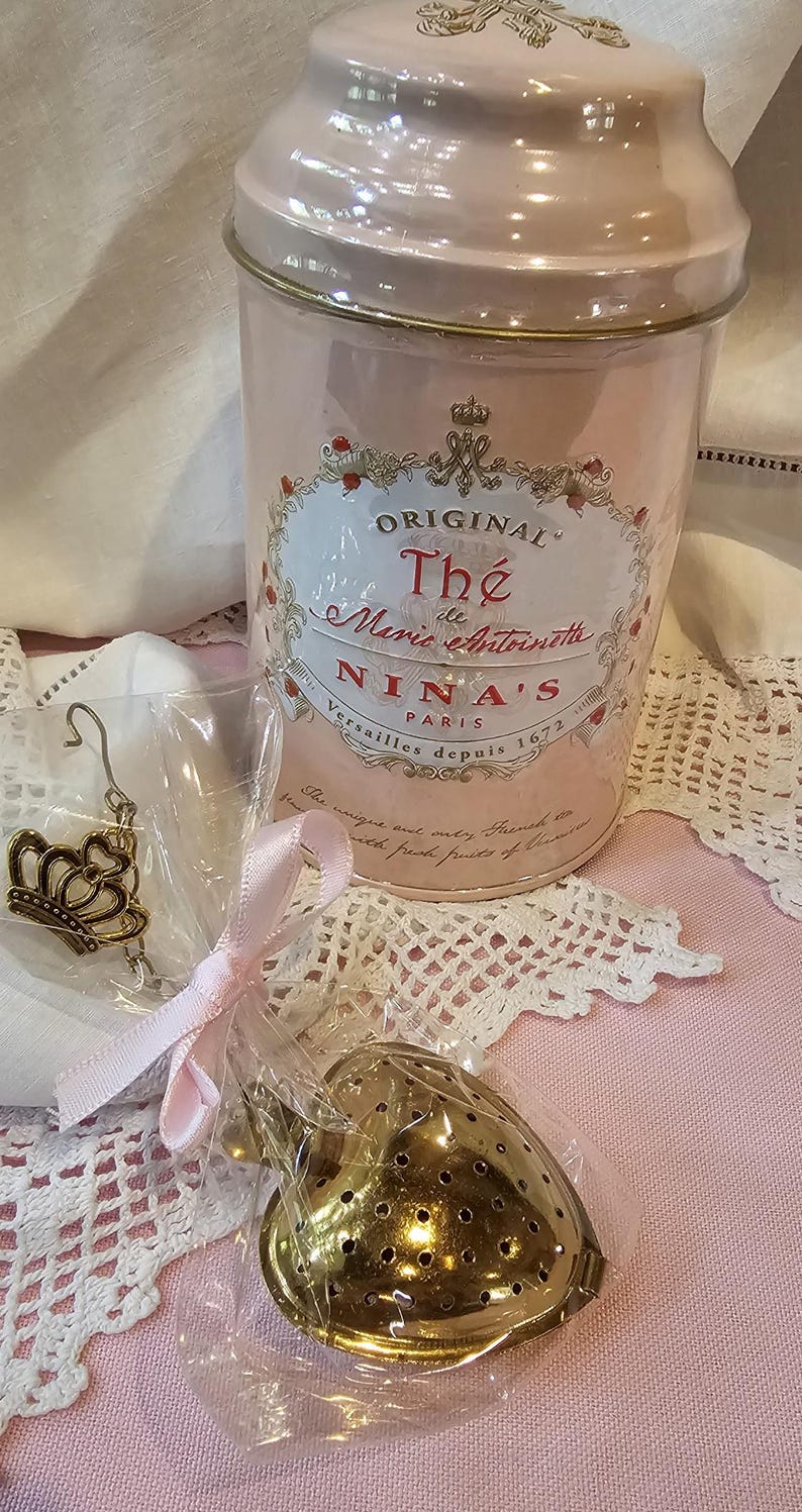 Op de afbeelding: Een roze theedoos met gouden rand en de tekst "ORIGINAL Th&eacute; de Marie Antoinette NINA'S PARIS Versailles depuis 1672". De doos staat op een witte kanttafelkleed. Een gouden hartvormige thee-ei is in een doorzichtige plastic zak met een roze lint.