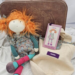 May include: A fabric doll with bright orange hair, wearing a floral dress, teal striped sleeves, and pink and teal leggings with purple shoes. Beside the doll is a beige canvas tote bag with a purple label reading Adèle and a matching product tag.