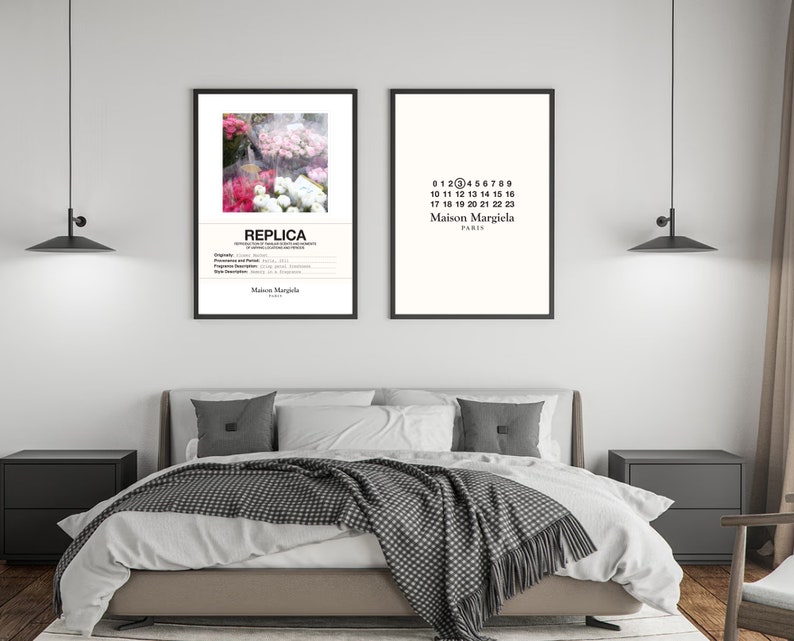 May include: Two black framed prints on a white wall above a bed with a white duvet and a grey and white blanket. The print on the left features a photograph of pink and white flowers with the text "REPLICA" and "Maison Margiela" below. The print on the right features the text "0123456789", "10 11 12 13 14 15 16", "17 18 19 20 21 22 23", "Maison Margiela", and "PARIS".