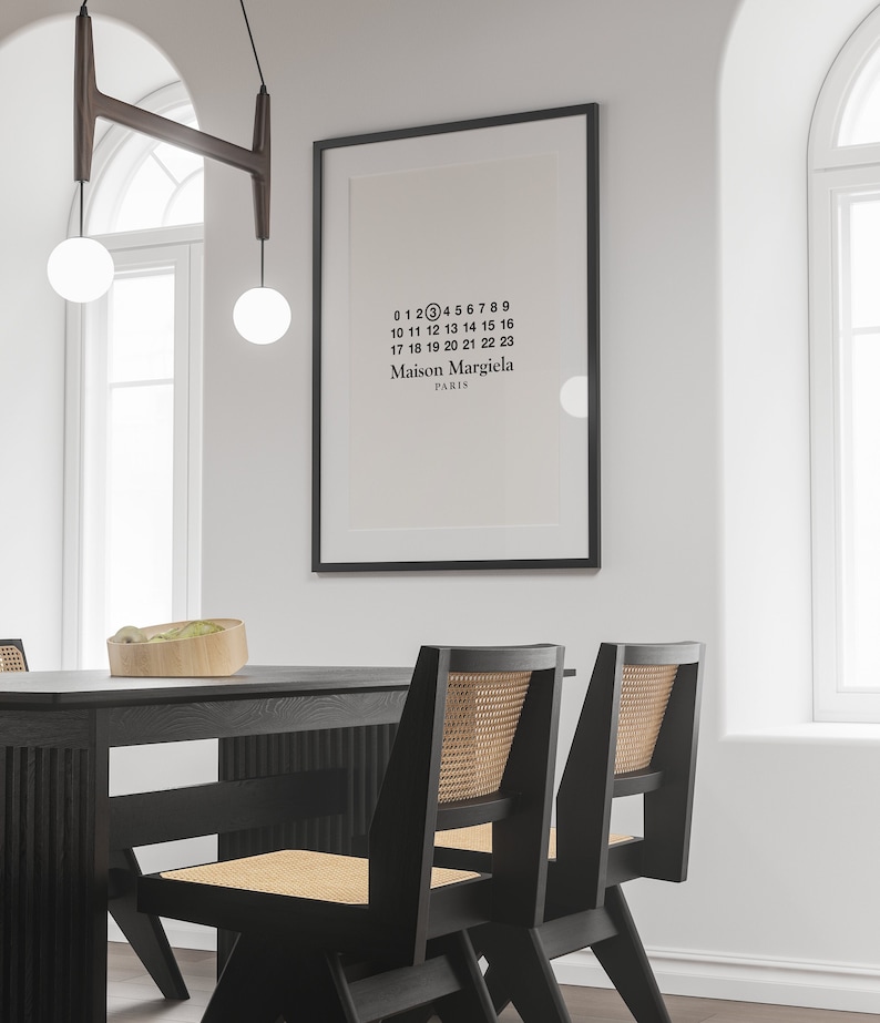 May include: A black framed poster with white text that reads "0123456789 10 11 12 13 14 15 16 17 18 19 20 21 22 23 Maison Margiela PARIS". The poster is hanging on a white wall above a black dining table with two black chairs.