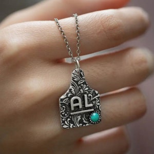 May include: Silver necklace with a silver pendant shaped like a livestock tag. The tag has a floral design and the initials "AL" engraved on it. A turquoise stone is set in the bottom of the tag.