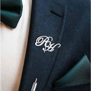 May include: A silver lapel pin with the initials "R & H" in a cursive script. The pin is on a dark blue suit jacket with a green pocket square.