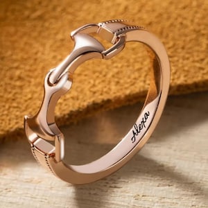 May include: A rose gold ring with a horse bit design. The ring is engraved with the name "Alexa".