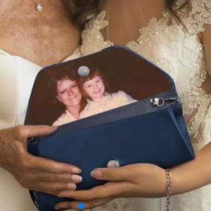 May include: A navy blue clutch with a silver chain strap. The interior of the clutch displays a photograph of two people. The clutch is held by a person wearing a white dress.