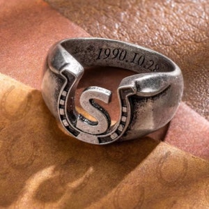 May include: A silver ring with a horseshoe design featuring the letter "S" inside. The ring is engraved with the date "1990.10.28".