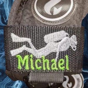 Puede incluir: Correa de buceo negra con un buceador bordado en blanco y el nombre "Michael" en verde.
