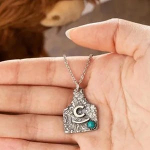 Peut inclure: Collier en argent avec un pendentif en argent en forme d'étiquette d'oreille de bétail. Le pendentif a un design orné et une pierre turquoise. Le pendentif est gravé de la lettre "C".
