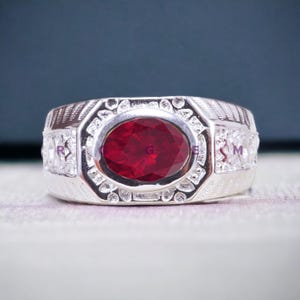 Peut inclure: Une bague en argent avec une grande pierre rouge ovale entourée de petites pierres blanches. La bague a un design détaillé avec une bande texturée.