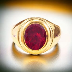 May include: A gold ring with a large, oval-shaped, red gemstone set in the center. The ring has a thick band and a raised bezel around the stone.