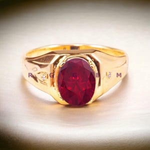May include: A gold ring featuring a large oval red gemstone and two smaller white gemstones set into the band.
