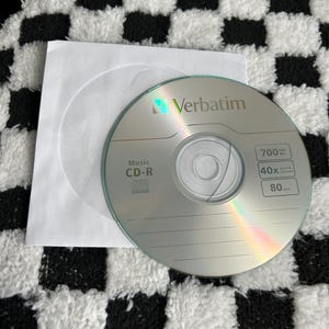May include: A silver Verbatim Music CD-R disc with a white paper sleeve. The disc has text indicating 700MB capacity, 40x speed, and 80 minutes of recording time. The background is a black and white checkered pattern.