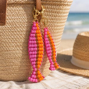 Puede incluir: Dos colgantes para bolso con forma de pez hechos con cuentas de color rosa vibrante y naranja brillante, unidos a un cierre de mosquetón de metal dorado. Los colgantes presentan un diseño bicolor vertical con una fila central de cuentas transparentes.