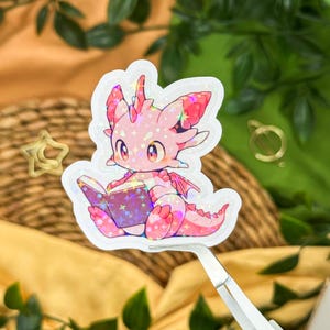 Könnte beinhalten: Ein holografischer Aufkleber mit einem niedlichen rosa Drachen, der ein Buch liest. Der Drache hat große Augen und kleine Flügel. Das Buch hat einen lila Einband mit Sternen. Der Aufkleber wird von einer Pinzette gehalten.