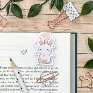 Può includere: Un segnalibro a forma di coniglio bianco con cuori rosa e uno sfondo marrone chiaro. Il segnalibro è appoggiato su un libro con il numero 12 sulla pagina.