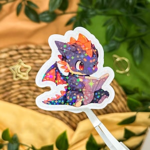 Könnte beinhalten: Holografischer Aufkleber eines Cartoon-Drachen, der ein Buch liest. Der Drache ist lila mit orangefarbenen Akzenten und roten Augen. Die Flügel sind mehrfarbig. Der Aufkleber hat einen weißen Rand.