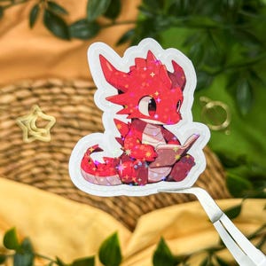 Könnte beinhalten: Ein holografischer Aufkleber mit einem roten Drachen, der ein Buch liest. Der Drache hat große Augen und sitzt. Der Aufkleber hat einen weißen Rand und wird von einer Pinzette gehalten. Der Aufkleber ist mit sternförmigen Glitzer verziert.