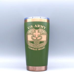 May include: Green insulated tumbler with a gold rim and base. The tumbler features the text "U.S. ARMY COMBAT MEDIC" with a gold emblem of a cross and snake. The tumbler has a clear plastic lid.