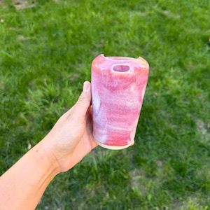 May include: A ceramic tumbler with a glossy, variegated glaze featuring shades of vibrant pink, soft white, and hints of orange. The vessel has a cylindrical shape with a slightly flared rim and a small, circular opening near the top edge.
