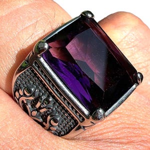 May include: A silver ring with a large, rectangular, deep purple gemstone. The ring has an ornate, textured design on the sides and is set against a light background.