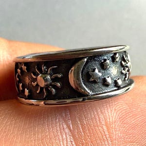 May include: A silver-toned metal ring featuring a dark, oxidized recessed background. The band is embossed with a repeating pattern of celestial motifs, including suns, crescent moons, and various small stars encircling the entire outer surface.