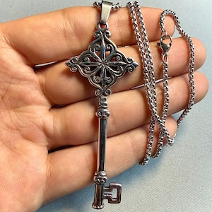 May include: A silver-toned metal pendant shaped like a decorative key with an ornate, filigree-style bow and a smooth, cylindrical shaft. The pendant is attached to a matching silver-toned box chain necklace with a lobster claw clasp closure.